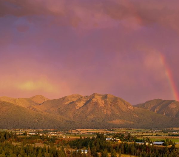 Enchanting aerial view capturing a rainbow's dance over Jocko Valley at sunset, framed by the mystical Mission Mountains